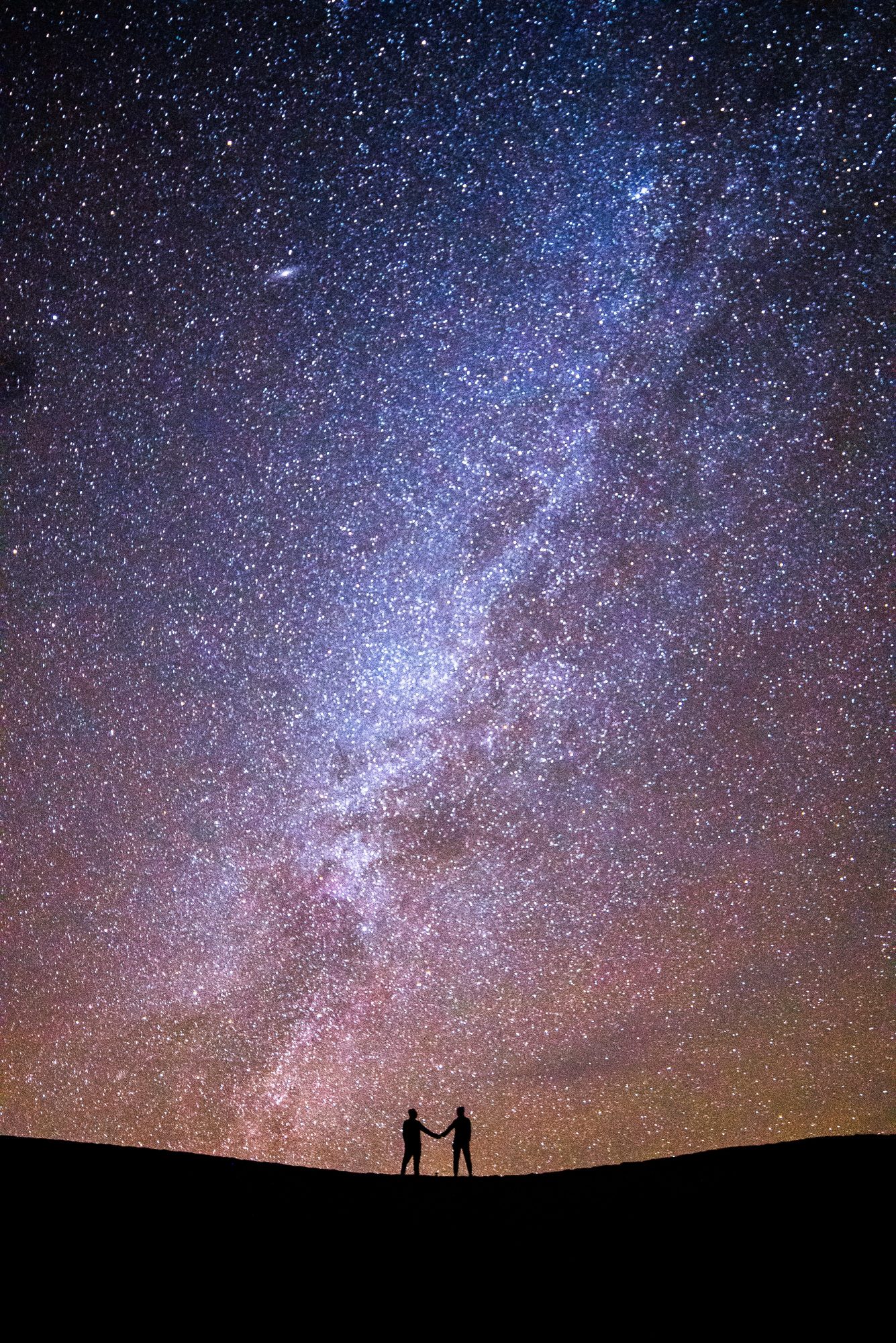 astrophotogphy long exposure photo of pj miles and joey ketterle holding hands under the milky way in the mesquite flat sand dunes in death valley national park in califronia | photo by jake landon schwartz